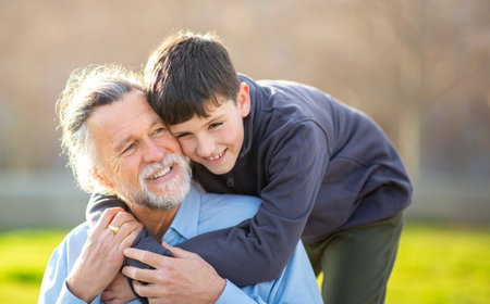 Close-up of a joyful older man smiling as his grandson embraces him from behind in a bright outdoor settingの写真素材