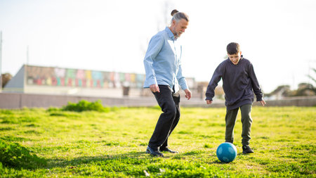 Smiling older man and boy share a fun moment kicking a blue soccer ball together on a sunlit grassy fieldの写真素材