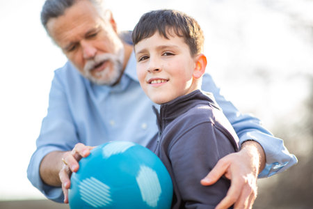 A smiling boy holds a blue ball with help from an older man, capturing a warm and supportive moment in the sunlight at a parkの写真素材