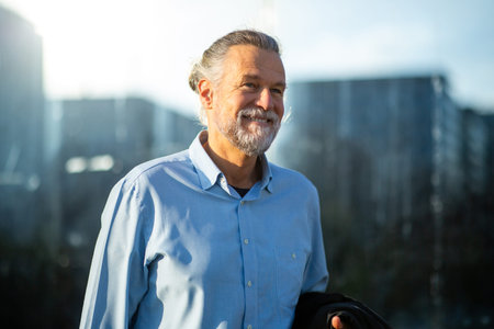 Portrait of a cheerful senior man in a light blue shirt enjoying a sunny day with a modern urban skyline in the backgroundの写真素材