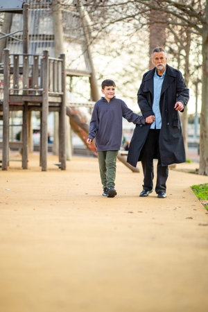 Grandparent and grandson share a warm moment as they walk hand in hand along a playground path surrounded by trees and play structuresの写真素材