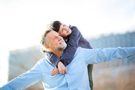 A cheerful boy clings to an older man from behind as they stretch their arms out playfully, imitating an airplane in a sunlit outdoor parkの写真素材