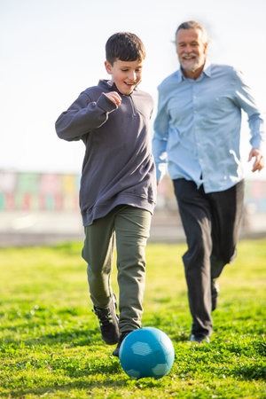 A smiling boy plays soccer with an older man in a sunny park, kicking a blue ball ahead as they both enjoy running togetherの写真素材