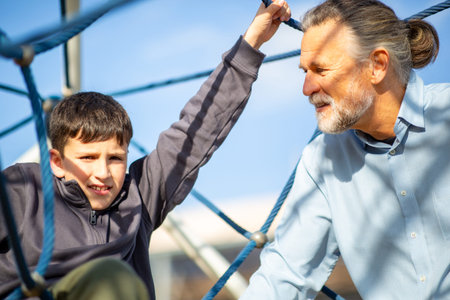 A young boy climbs a rope web at a sunny outdoor playground, supported by an attentive older man who smiles in encouragementの写真素材