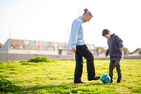 Elderly man and young boy enjoy an active moment kicking a blue soccer ball together on a sunny green fieldの写真素材
