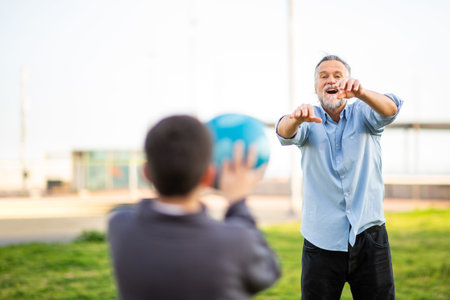 A cheerful grandfather reaches out to catch a ball tossed by his grandson during an energetic outdoor play session on a grassy fieldの写真素材
