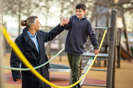 Older man in a dark coat holding a boy's hand while helping him balance on a yellow rope in an outdoor playgroundの写真素材