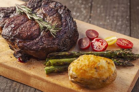 a bone-in tomahawk ribeye steak meal with twice baked potato, asparagus, tomatoes and sprig of rosemary on a wood plank over wooden background, angled viewの写真素材