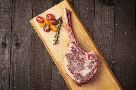 a bone-in tomahawk ribeye steak being prepared for grilling with tomatoes, seasoned with salt, pepper and a sprig of rosemary on a wood plank over wooden background, overhead viewの写真素材