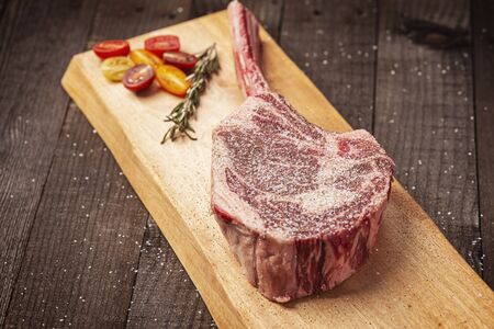 a bone-in tomahawk ribeye steak being prepared for grilling with tomatoes, seasoned with salt and papper and a sprig of rosemary on a wood plank over wooden background, angled viewの写真素材