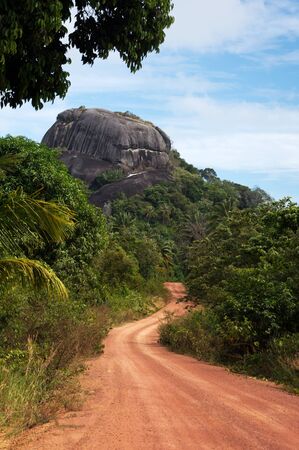 Dusty road going through green colored bushes leading to a big black rock at midday and nobody around on the island Belitung, Indonesia.の写真素材