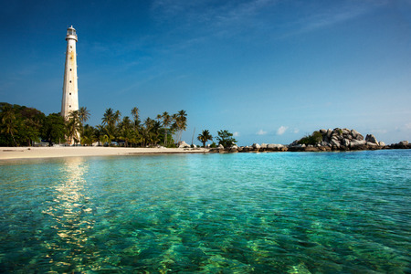 White lighthouse standing on an island in Belitung at daytime surrounded by clear blue green colored ocean water with no people around.の写真素材