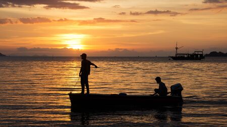 Silhouette of two fishermen on a small fishing boat with outboard motor on the ocean at nightfall with an orange sunset at the background in asia.の写真素材