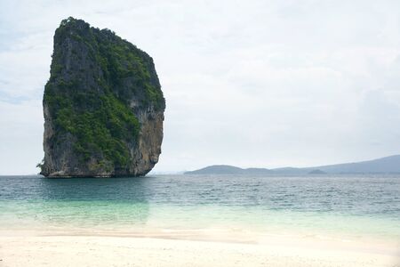 Big high rock cliff filled with green vegetation surrounded by turquoise blue colored ocean water next to a tropical white sand beach with horizon view at midday, Krabi Thailand.の写真素材