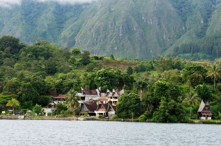 Several houses build at the foot of a mountain next to ta lake in Sumatra Samosir Island in the afternoon, Indonesia.の写真素材