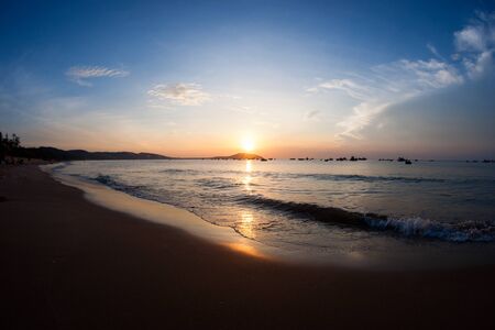 Silhouette of several small fishing boats at sea seen from sandy beach in the morning during golden colored sunrise with blue sky, Vietnam Asia.の写真素材