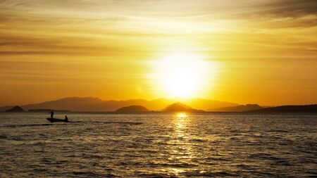 Silhouette of two fishermen on a small fishing boat with outboard motor on the ocean at nightfall with an orange sunset at the background in asia.の写真素材