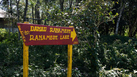 Red sign next to green vegetation giving easy direction with yellow letters and arrow towards ranamese lake danau, Indonesia.の写真素材