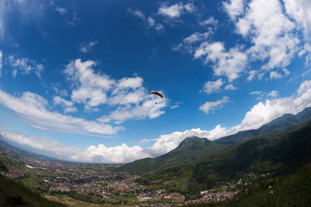 One paraglider in the blue sky with red parachute gliding for fun and excitement towards the mountains, villages and fields on the ground.の写真素材