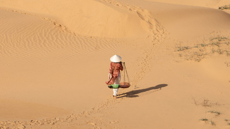 Asian woman carrying two baskets attached to a wooden stick on her shoulders in the desert at noon and heading home.の写真素材