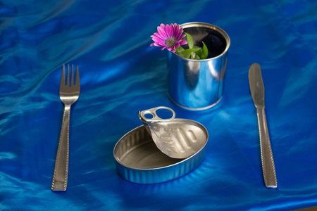 Empty sardine tin can with nothing to eat together a fork, knife and pot with a purple flower and green leaves on the side on blue Backgroundの写真素材