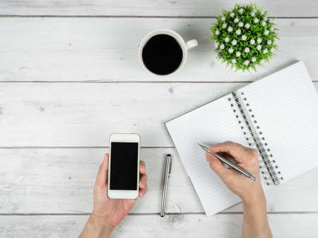 Workspace on white wood table top view copy space,Hand hold silver pen and smartphone in hand with notebook on the deskの写真素材