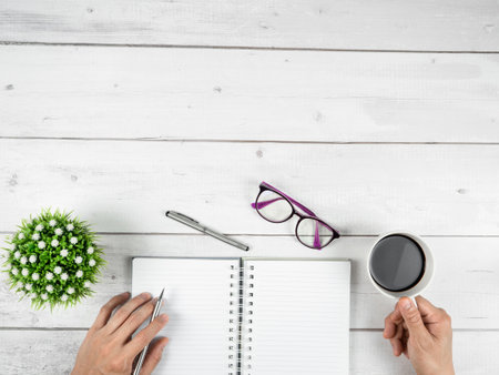 Office workspace top view copy space ,Hand with silver pen and coffee cup and glasses and blank page notebook on white wood tableの写真素材