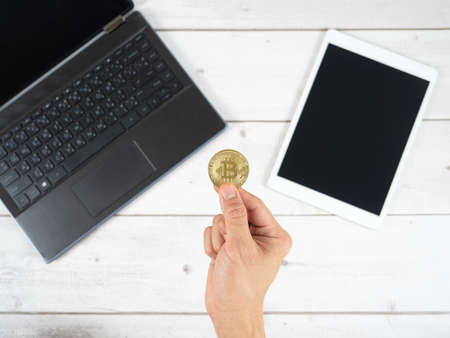 Hand holding gold bitcoin with laptop and tablet on table background top viewの写真素材