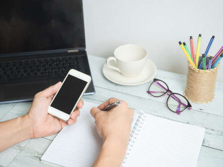 Hand holding smartphone and writing on notebook workspace on white wood table backgroundの写真素材