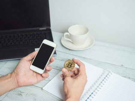 Hand holding gold bitcoin and mobilephone with workspace on white wood table backgroundの写真素材