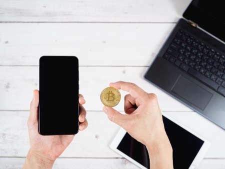 Hand holding gold bitcoin and mobilephone with laptop and tablet on wood table backgroundの写真素材