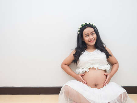 Asian pregnant woman sitting on the floor touch her belly with happy smile face on white background