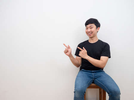Asian man sitting on chair with smile face feel happy and cheerful point finger at copy space on white backgroundの写真素材