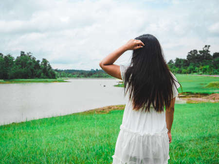 Asian girl stand with river and forest and mountain fresh nature in vacation,Long hair,Backside Woman white dress in green grass filed and lake tree cloudy of sky landscape backgroundの写真素材