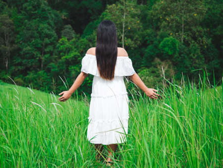 Woman in white dress stand in meadow with forest,mountain background,Travel woman in grass field with tree background beautiful nature vacationの写真素材