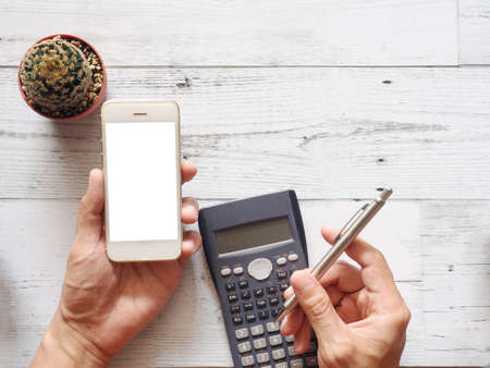 Hand holding cellphone white screen and using calculator with cactus and silver pen on white wood table nature shadow and sunlight top view and space business conceptの写真素材