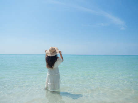 Woman white dress catch hat stand in sea water blue ocean blue sky landscape background beautiful beach travel at holiday conceptの写真素材