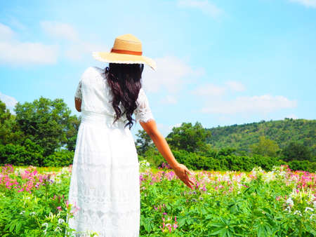 Young woman traveler white dress standing in flower garden and touch pink flower with valley and blue sky background landscapeの写真素材