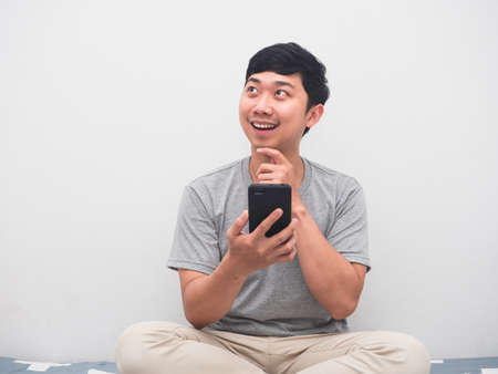 Young positive man sitting on the bed holding smartphone thinking with shopping
