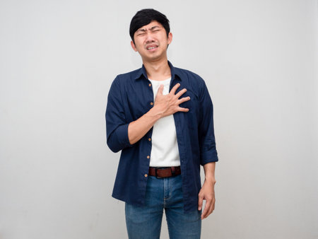 Negative asian man stand gesture hold his chest feels pain his heart isolatedの写真素材