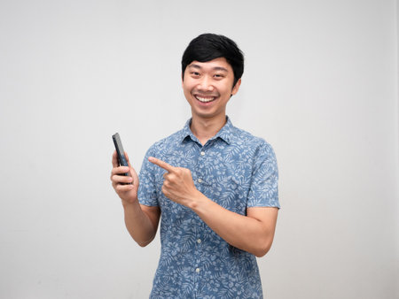 Cheerful asian man smile and point finger at mobile phone in hand isolatedの写真素材