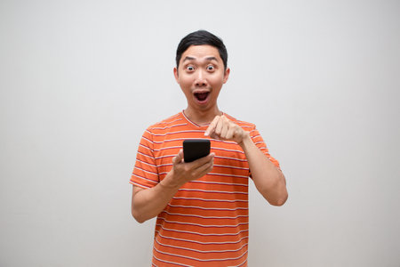 Asian man orange shirt feels excited point finger at moblie phone isolatedの写真素材