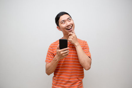 Asian man orange shirt holding smartphone gesture thinking looking up isolatedの写真素材