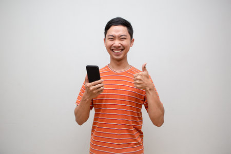 Asian man orange shirt feels happy holding mobile phone and show thumb up isolatedの写真素材