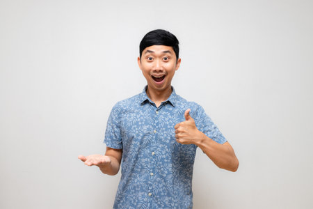 Positive asian man blue shirt amazed show thumb up with empty hand up isolatedの写真素材