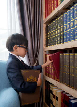 Little boys in suit looking and choose book in shelf.