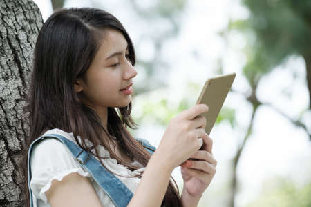 Young Asian teenage girl learning online with tablet and notebook in park.