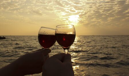 two glasses of red wine in hand. Romantic day by the sea at sunset. The concept of wedding or Valentine's Day celebrations.の写真素材