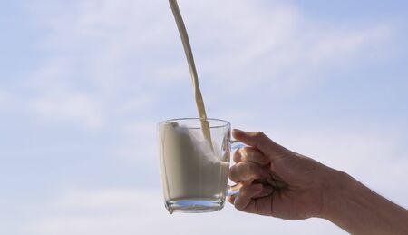 milk pours into the cup. hand holding a glass glass with dairy product against a clear sky.