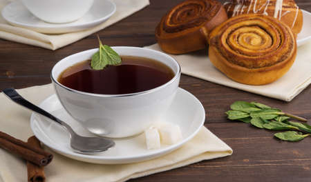 hot black tea with mint leaves in a white mug on a wooden table. selective focus. morning tea ceremony. aromatic drink with sugar and mint. delicious cinnamon rolls for breakfast. meal conceptの写真素材
